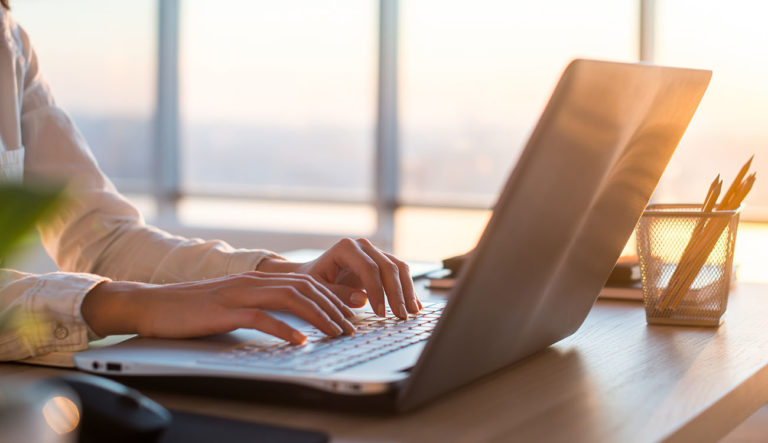 Eine Person tippt auf einem Laptop an einem Schreibtisch mit Blick auf große Fenster im Abendlicht. Rechts im Bild steht ein Becher mit Bleistiften. Die warme Lichtstimmung vermittelt Ruhe, Verlässlichkeit und konzentriertes Arbeiten – Werte, die bei Nesari im Mittelpunkt stehen.